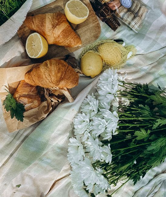 A still life scene featuring two golden-brown croissants wrapped in brown paper, garnished with sprigs of fresh green parsley, accompanied by two lemon halves placed on a piece of parchment paper. Adj