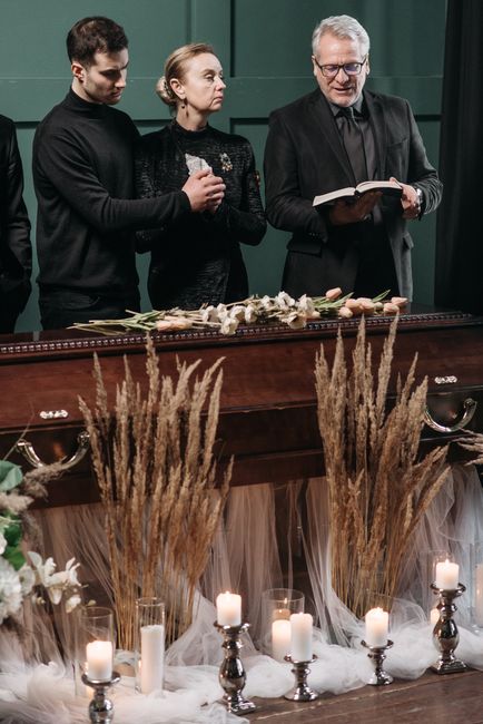 An indoor funeral or memorial setting featuring a dark wooden casket decorated with white and cream flowers, including roses and lilies, arranged on top. In front of the casket, there are tall arrange
