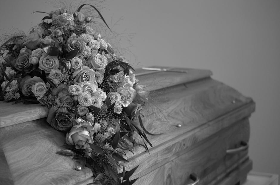A large floral arrangement featuring an assortment of fresh roses in various stages of bloom, predominantly in light and dark shades, resting on the surface of a wooden casket. The bouquet includes ro