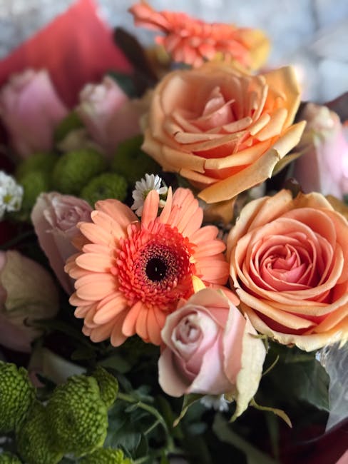 A close-up of a floral arrangement featuring peach and pink roses with softly curled petals, accented by a peach gerbera daisy with a dark central disc, surrounded by small white and lavender buds. Th